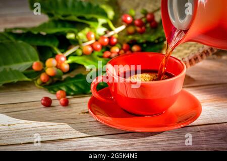 Coffee being poured into red cup with coffee seeds and leaves over wooden table. Stock Photo