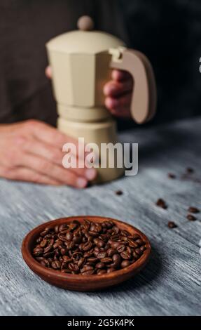 a young caucasian man is preparing some coffee in a beige moka pot, sitting at a gray rustic wooden table and some roasted coffee beans in the foregro Stock Photo