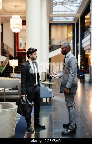 Vertical side view portrait of two successful businessmen discussing work while standing in hotel lobby Stock Photo