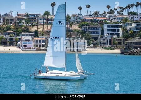 A white trimaran sails out of the channel at Newport Beach California and into the Pacific Ocean with exclusive homes in the background Stock Photo