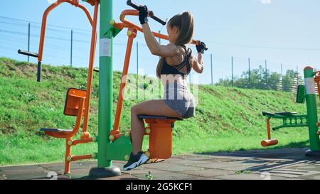 Side view of fit woman practicing wide grip lat pulldowns using simulator at sports ground. Stunning girl with muscular body wearing sports outfit training back outdoors in sunny summer morning. Stock Photo
