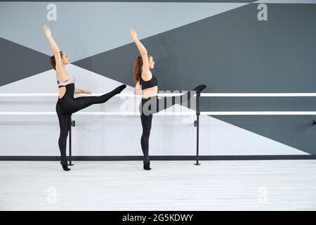 Back view of adult female instructor and young teenager in black sportswear learning choreographic move, holding handrail. Two synchronized women practicing stretching in hall. Gymnastics concept. Stock Photo