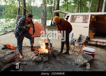 Two men cooking food together on a campfire during camping in the forest Stock Photo