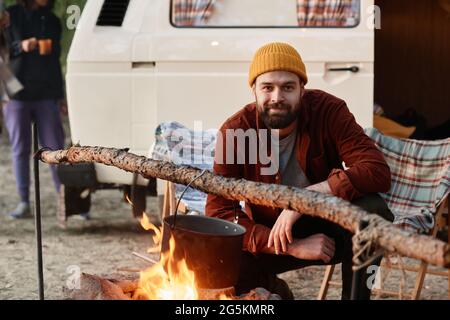 Portrait of young bearded man looking at camera while cooking food on a fire during camping in the forest Stock Photo