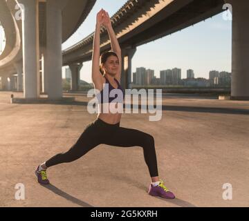 Fit jogger lunging with raised arms during fitness training under bridge in city Stock Photo
