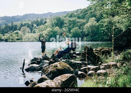 woman sitting on rocking chair by a lake whilst her kids throw rocks Stock Photo