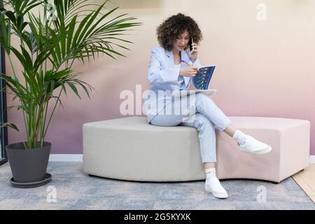 Happy female office worker sitting in a couch talking on the phone and taking notes. Stock Photo
