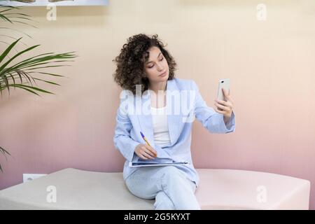 Happy female office worker sitting in a couch smiling and taking a selfie with her phone. Stock Photo