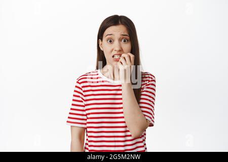 Nervous brunette 20s girl, biting finger nails and looking worried, watching smth scary, panicking or being anxious, standing against white background Stock Photo