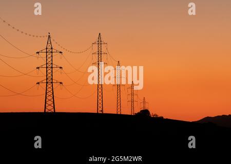 Towers of electricity over mountains at sunset. Silhouettes of power lines and towers with a stork on top of one of the towers. Stock Photo