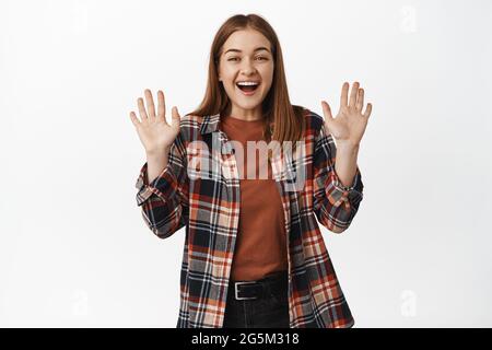 Image of beautiful girl smiling friendly, waving hands to say hello, hi gesture, welcome or greet someone, goodbye wave, standing against white Stock Photo