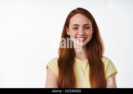 Portrait of young redhead girl face, long natural foxy hair and white smile, looking happy and carefree, standing relaxed in t-shirt against white Stock Photo
