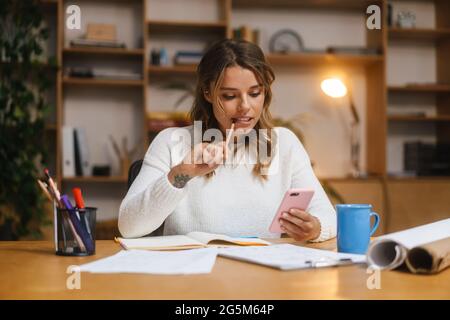 Smiling attractive young woman entrepreneur using mobile phone while working with documents at the office desk Stock Photo