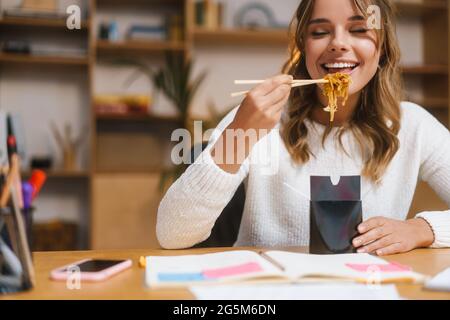 Smiling young woman entrepreneur having lunch while sitting at the office desk, eating noodles Stock Photo
