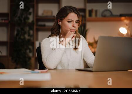 Serious businesswoman in earphones watching webinar on laptop making notes, learning studying computer course, making call, participating online confe Stock Photo