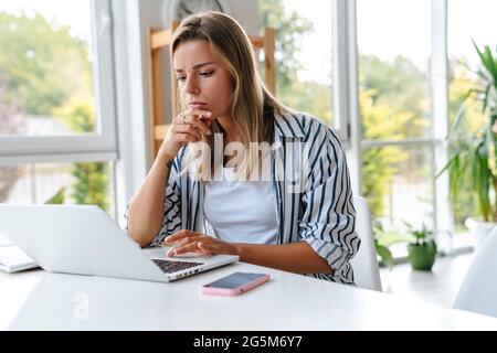 Attractive pensive confident young woman working at creative office, using laptop computer Stock Photo