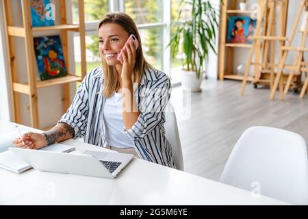 Smiling attractive young woman creative designer using mobile phone while working at the studio Stock Photo
