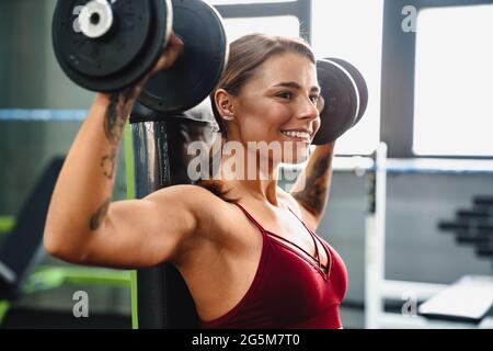 Image of cheerful young woman fitness coach making sport exercise for arms with dumbbells in gym Stock Photo