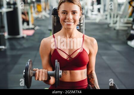 Image of cheerful young woman fitness coach making sport exercise for arms with dumbbells in gym Stock Photo