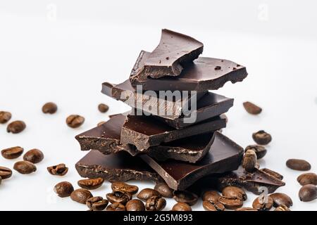 A stack of dark cracked chocolate and black coffee beans on a white background. Pieces of bitter chocolate. Food photography. Close-up. Stock Photo