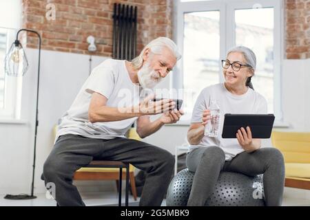 Smiling mature man and woman taking break during morning exercises and using modern tablet with smartphone. Concept of people, recreation and gadgets. Stock Photo