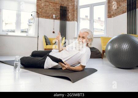 Grey haired man dressed in sport clothes using digital tablet while workout on yoga mat. Bearded male looking at camera and showing thumb up. Stock Photo