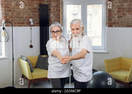 Energetic retired couple in casual clothes moving actively at bright living room. Two happy people dancing together at home. Leisure time concept. Stock Photo