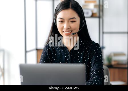 Positive asian young woman, with headset, call center worker or support service operator, sitting at table, using laptop, advising clients, communicate online, smiling friendly Stock Photo