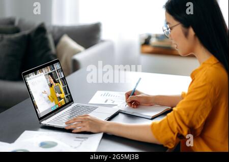 Asian female student watching lesson online and studying from home. Japanese or chinese girl taking notes while looking at laptop screen, listening lecture from female teacher on video call Stock Photo