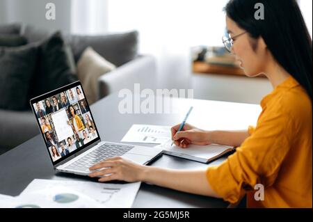Side view at asian female student watching lesson online and studying distantly. Young woman takes notes, listening lecture from female teacher on video call.Video conference with teacher and students Stock Photo