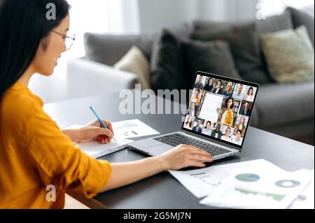 Video conference with teacher and students. Clever asian female student watching lesson online and studying distantly. Young woman takes notes, listening lecture from female teacher on video call Stock Photo