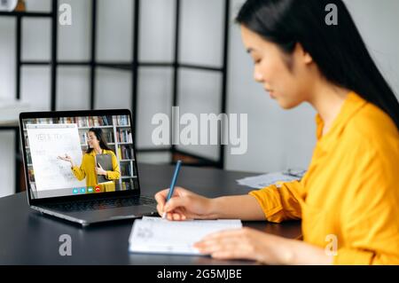 Clever asian female student gains knowledge watching lesson online and studying distantly. Young woman takes notes during webinar, listening lecture from female teacher on video call. Online education Stock Photo