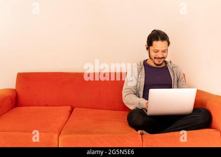 Young man working using computer laptop at home in the living room sitting on an orange couch sofa at home. Stock Photo