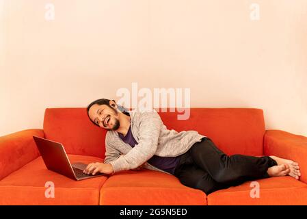 Young man dating online using the computer laptop laying on an orange couch sofa at home in the living room. Stock Photo