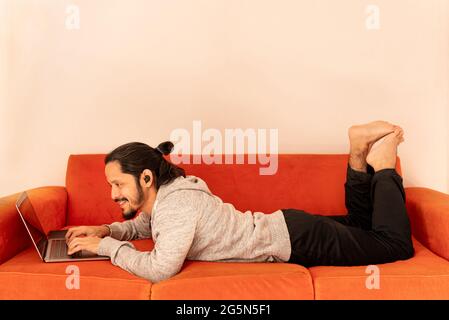 Young man dating online using the computer laptop laying on an orange couch sofa at home in the living room. Stock Photo