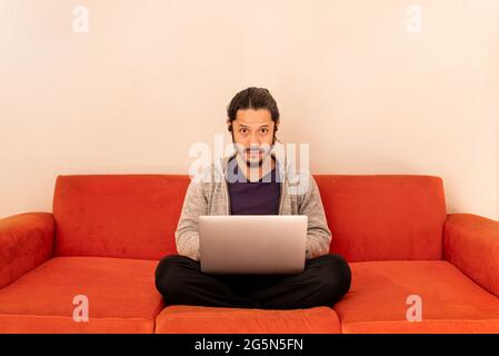 Young man working using computer laptop at home in the living room sitting on an orange couch sofa at home. Stock Photo