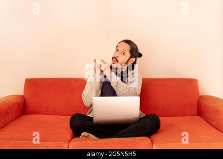 Young man pointing up with finger sitting on an orange couch sofa in the living room at home. Concept for sales time. Stock Photo