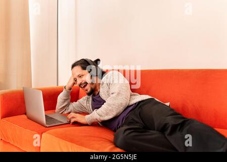 Young man working using computer laptop at home in the living room sitting on an orange couch sofa at home. Stock Photo