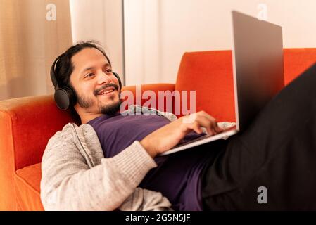 Young man working using computer laptop at home in the living room sitting on an orange couch sofa at home. Stock Photo