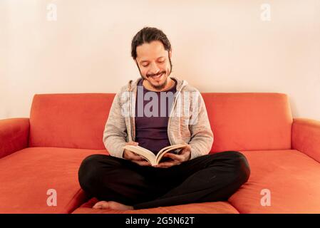 Young latin man reading a book on a sofa in the living room at home Stock Photo