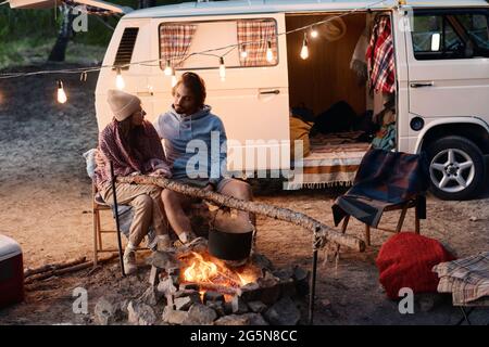 Young couple talking and resting near the fire during camping in the forest Stock Photo