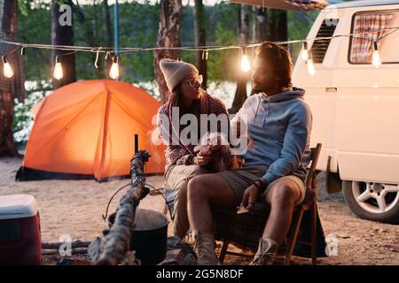 Young couple talking to each other during their camping in the forest with tent and van in the background Stock Photo