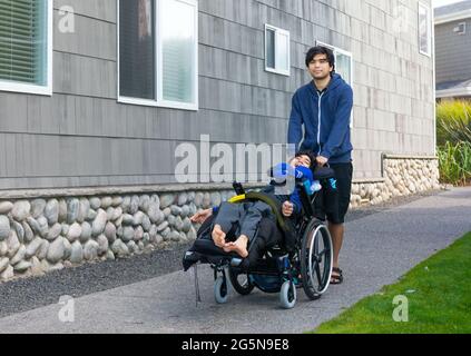 Biracial young man pushing disabled little boy in wheelchair on sidewalk next to building outdoors Stock Photo
