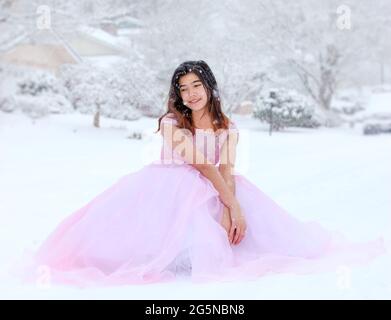 Biracial teen girl in long formal pink gown sitting outdoors in beautiful snowfall Stock Photo