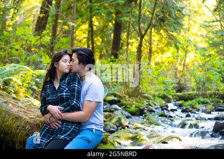 Biracial young couple sitting along rocky stream in green woods hugging Stock Photo