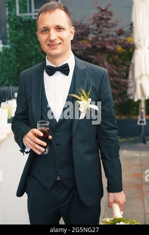 Handsome groom in formal suit having drink and holding bride's bouquet Stock Photo