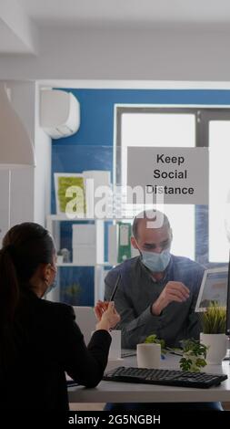 Business people working at financial graphs using digital tablet while sitting at office desk in corporate company. Team wearing face masks maintaining social distancing to avoid coronavirus pandemic Stock Photo