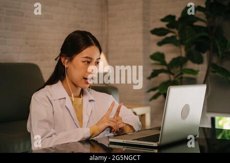 Successful young Asian female doctor with labcoat and earphones doing video conference call for consultation with patient from home at late night Stock Photo
