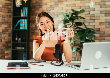 Portrait of cheerful and successful young female Asian influencer promoting products while creating podcast using mic and laptop at home Stock Photo