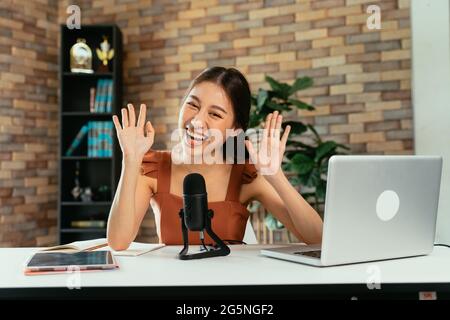 Portrait of cheerful young Asian female blogger using mic and laptop with digital tablet to interview guest during live stream at home Stock Photo